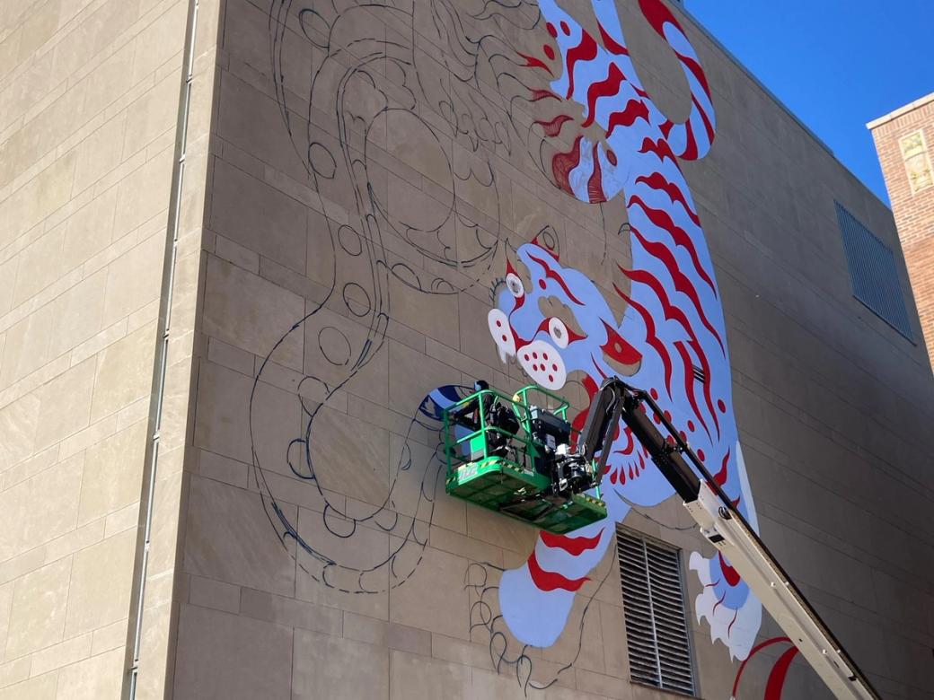 A large mural of a white tiger with red stripes, partially painted, in progress on the tan stone exterior of a building. A green boom lift with two workers is raised alongside the wall, working on the upper portion.