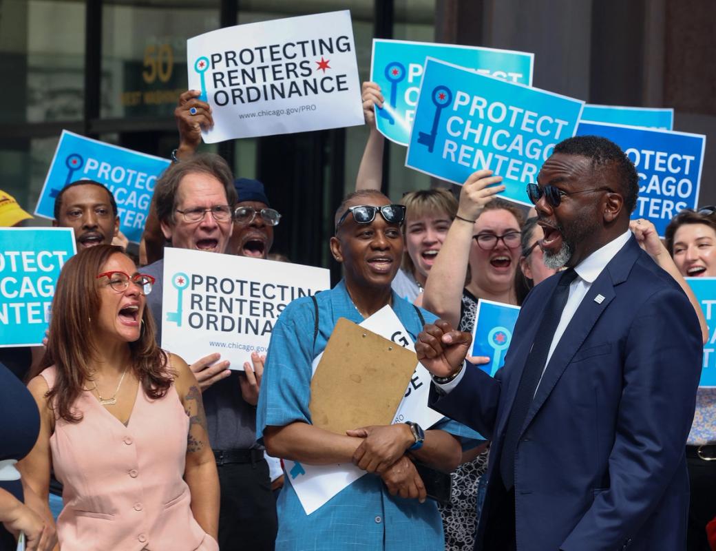 A group of people on stage. In the front there's a woman wearing pink, a man with a clipboard in the middle and a man in a suit on the right. In the back there are people holding signs that say "protecting renters ordinance" and "protect chicago renters"