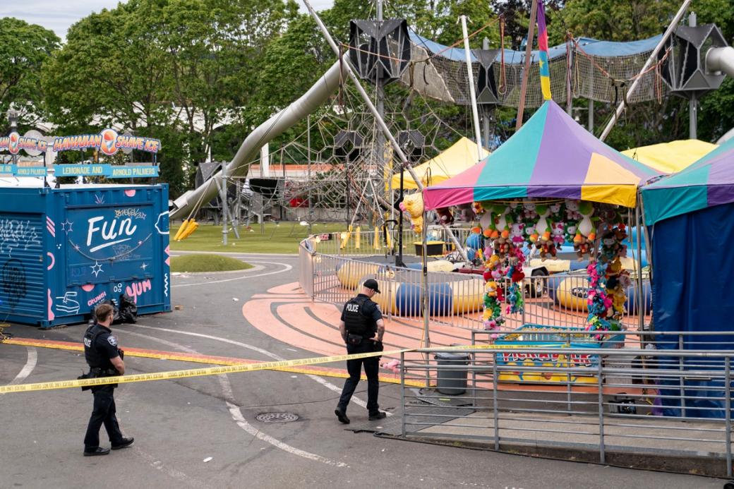 Two police officers walk through a caution taped off crime scene at Seattle Center with pastel colored tents and a booth reading “fun” on either side of them. The grounds are otherwise empty.