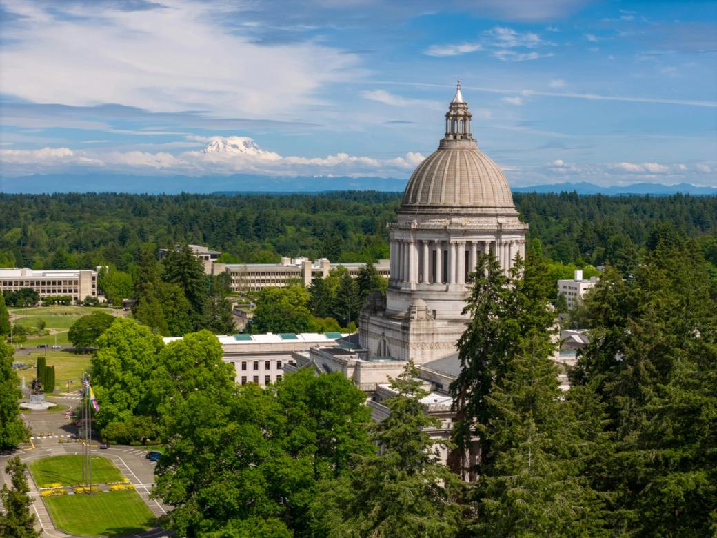 The olympia capitol dome, surrounded by green trees, with Tahoma and a blue cloudy sky visible in the background