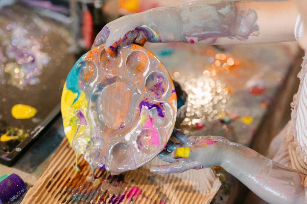 A child with paint-splattered arms holds a paint palate covered in colors.