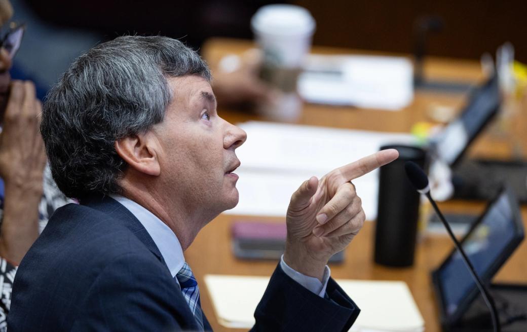 A man sitting at a desk with a microphone and pointing his finger off camera