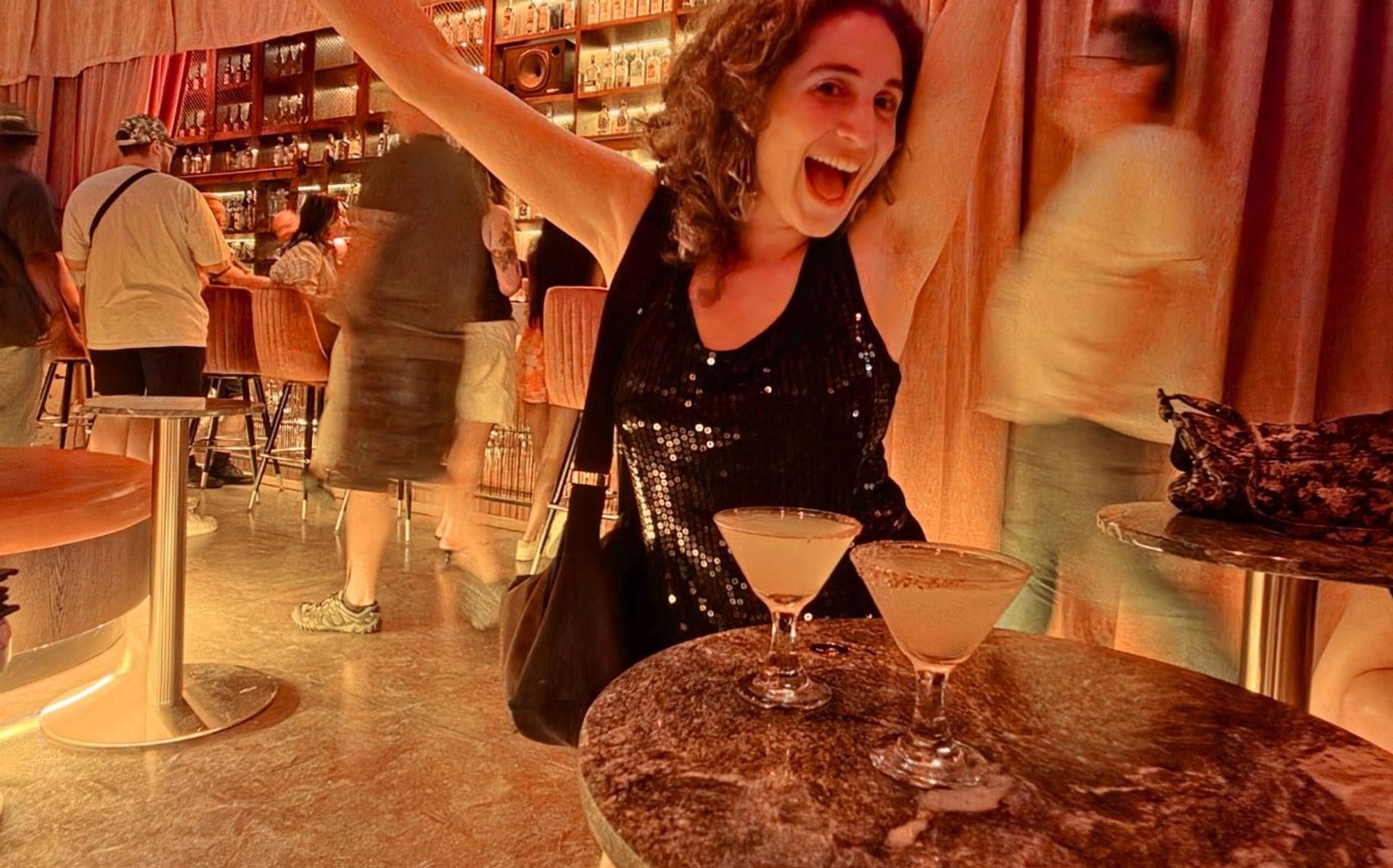 A woman with her arms up at a bar with two drinks in front of her