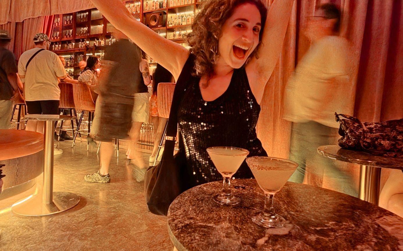 A woman with her arms up at a bar with two drinks in front of her