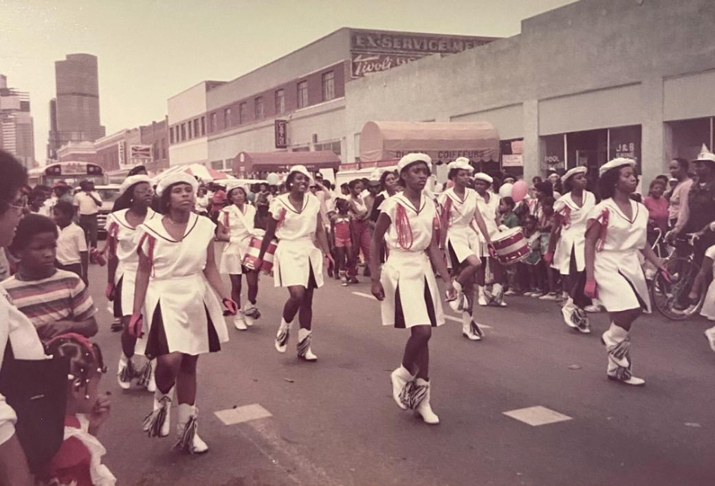 A vintage photograph of a marching drill team of young Black women in white sailor-style uniforms and white boots performing in the Juneteenth street parade, with crowds lining the sidewalks and city buildings visible in the background.