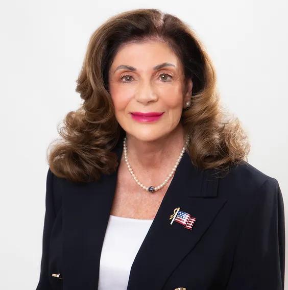 Headshot of a pale woman with shoulder-length curly hair smiling at the camera. She wears a dark suit with an American flag pin and a pearl necklace.