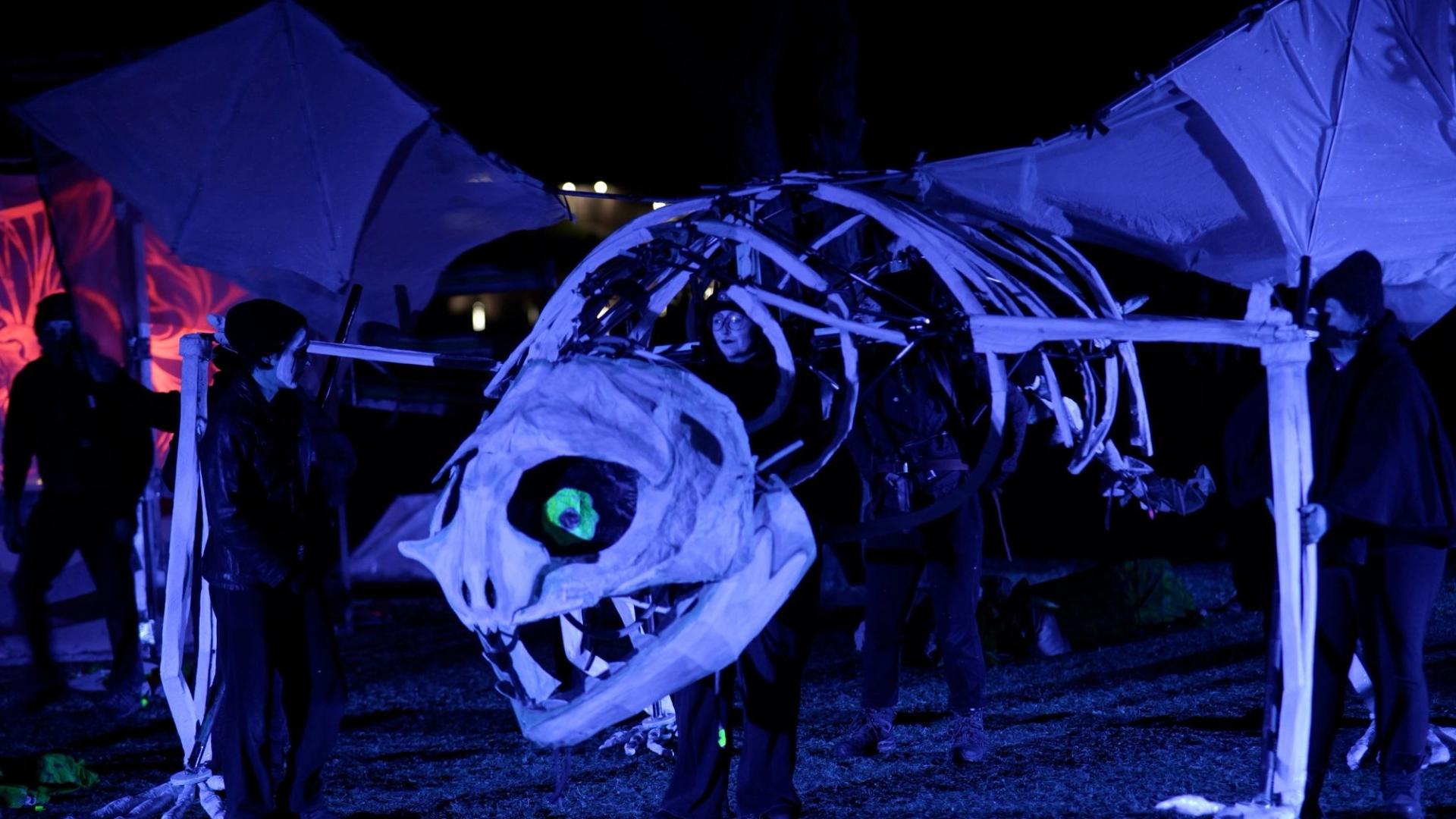 A puppet of a giant lizard skeleton is operated by volunteers wearing all black at night. Multiple people stand inside the ribcage to help the structure move, and one person stands by each leg to move them as if the creature is walking. 