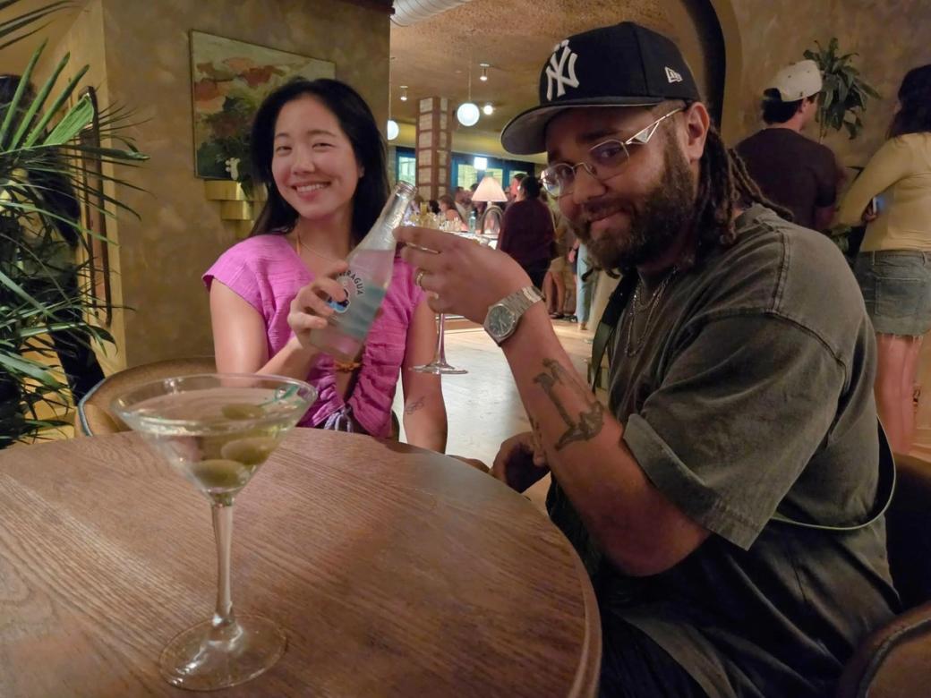 Two people at a table holding up their drinks. On the table there's a martini with olives.