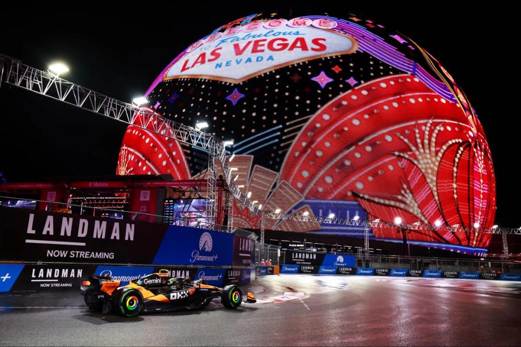 A McLaren MCL39 Mercedes racecar driving on the Strip Circuit, with the Sphere in the background lit up in red, gold, purple, and other colors, with the words "Welcome to Fabulous Las Vegas Nevada"