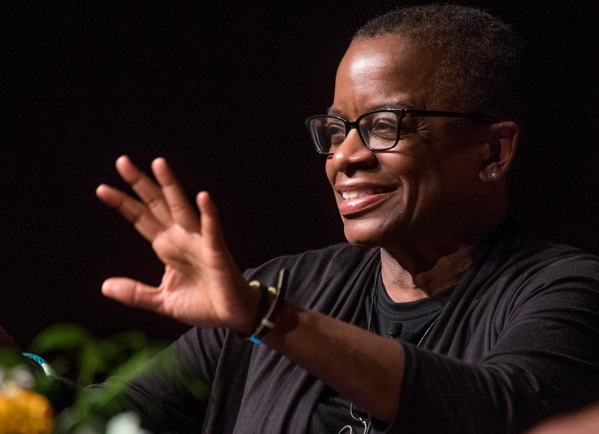 A woman wearing black-rimmed glasses and smiling with her left hand outstretched.