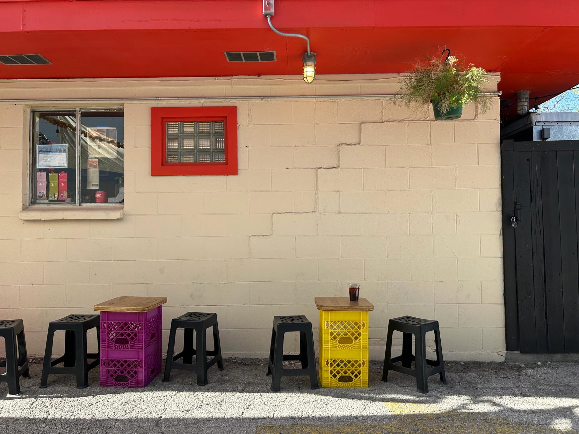 The facade of a white building with a red roof. Small tables made of milk crates are accompanied by small black stools.