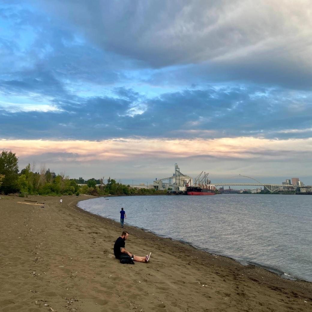 A river beachfront at sunset on a cloudy day with a bridge, an industrial plant, and a large ship in the distance, and two people on the shore.