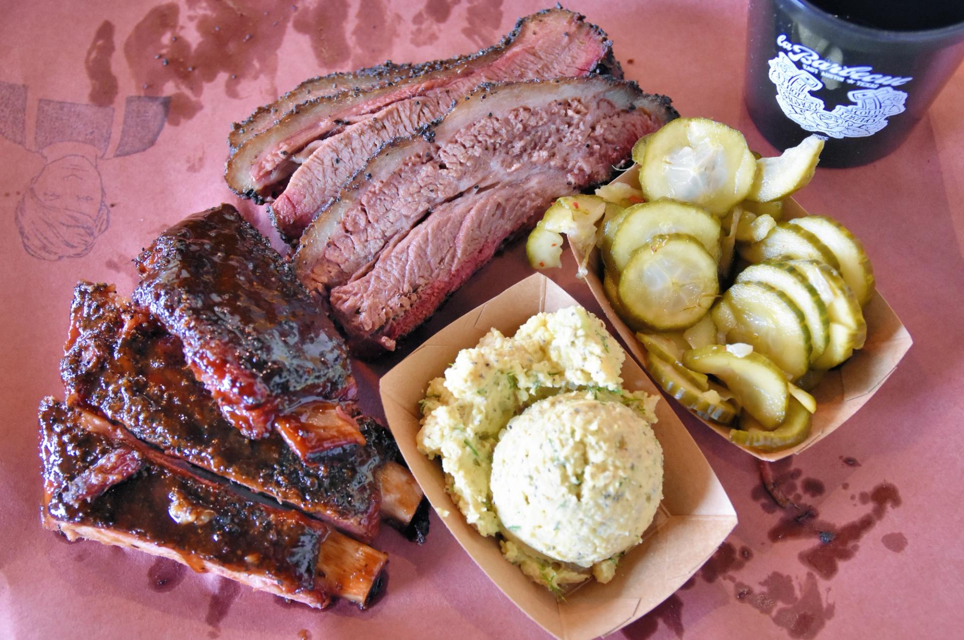 Slices of brisket, ribs, a basket of pickles and potato salad sit on a sheet of brown paper.