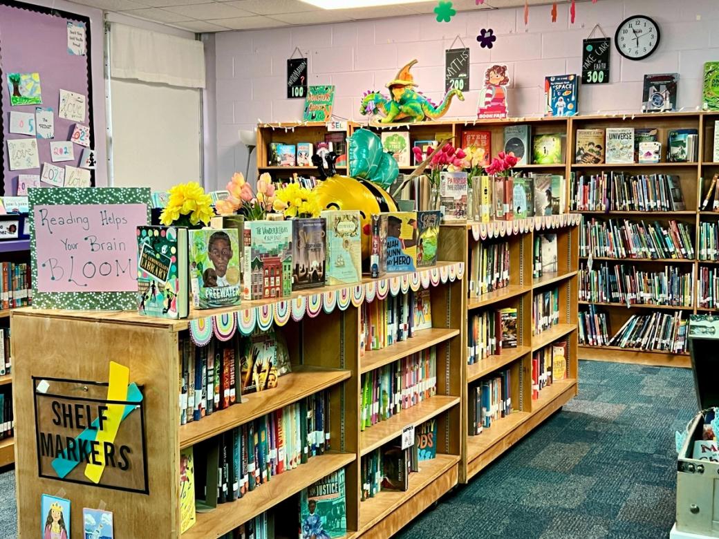 Light brown bookshelves, both short and tall, packed with children's books. Books, flowers, and colorful decorations are on top of the shelves, as well as a sign that says "Reading helps your brain bloom." The carpet is teal, and the walls purple.