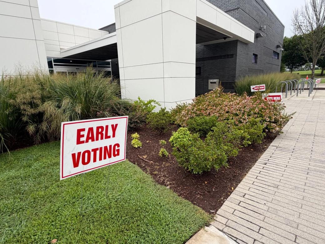 A white sign that says "Early Voting" in red letters on a grass plot in front of a white and gray building. More election signs can be seen in the background.