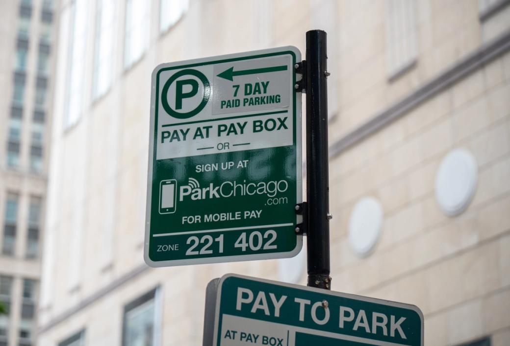 A green and white street parking sign on a black pole, indicating seven-day paid parking and payment by pay box or mobile app. The sign lists ParkChicago.com and zone numbers 221 and 402. A second parking sign is partially visible below.