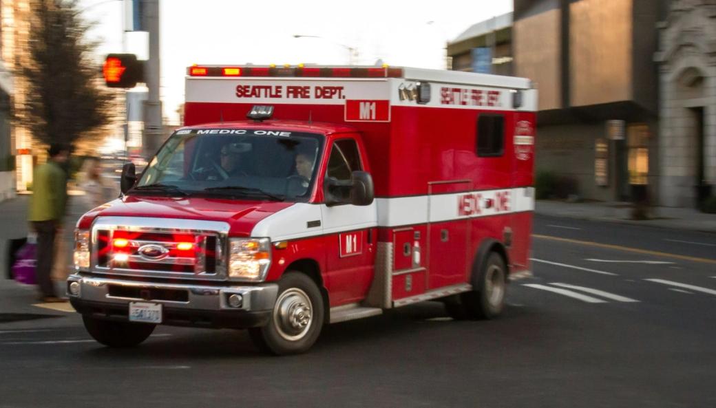 A red and white Seattle Fire Dept. truck turns a corner across an intersection with its lights flashing and motion blur behind it in the image.