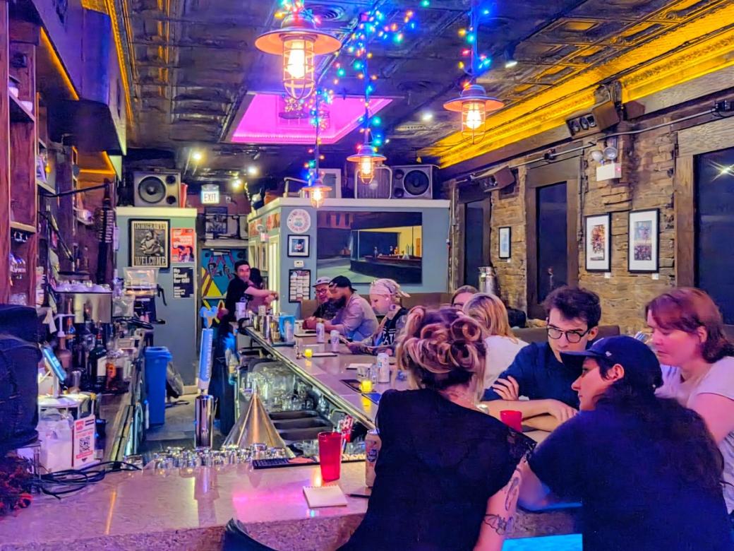 The inside of a bar with people gathered around the bar top It is fairly well lit but the lighting is colorful