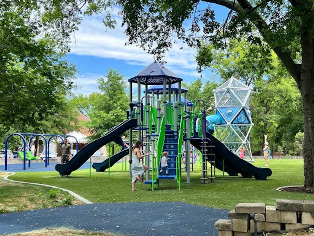 Nashville's Fannie Mae Dees Park on a sunny day. A jungle gym sits on green grass with green staircases and black slides. A toddler stands on a stair.