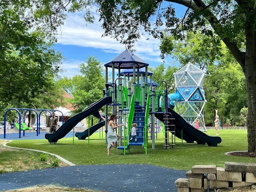 Nashville's Fannie Mae Dees Park on a sunny day. A jungle gym sits on green grass with green staircases and black slides. A toddler stands on a stair.