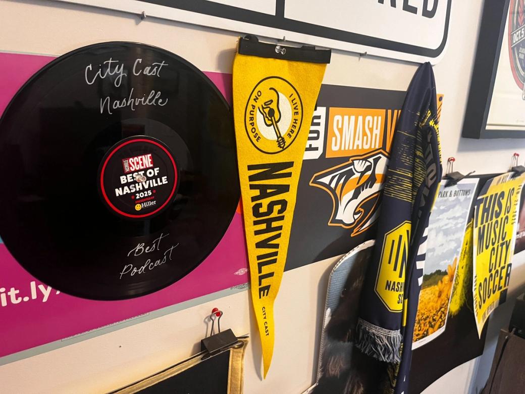 A yellow pennant that says, "I live here on purpose. Nashville City Cast" in black letters. It hangs from a wall surrounded by other Nashville memorabilia: A record that says Best of Nashville, a Nashville Predators poster, and a Nashville SC flag.