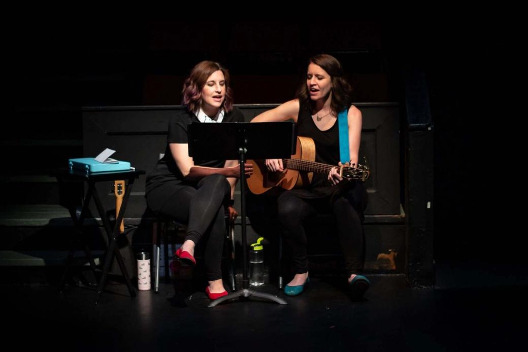 Two women sit on chairs on a darkened stage with light on them, one holding a guitar.