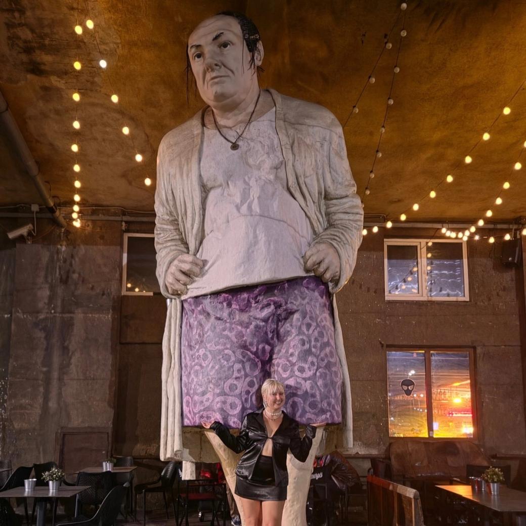 A large hand-painted statue of a man wearing a cardigan, apron and purple patterned shorts stands inside a dimly lit restaurant or bar. A woman in a black outfit stands in front of the statue, posing with her arms raised. String lights hang from the ceiling and walls.