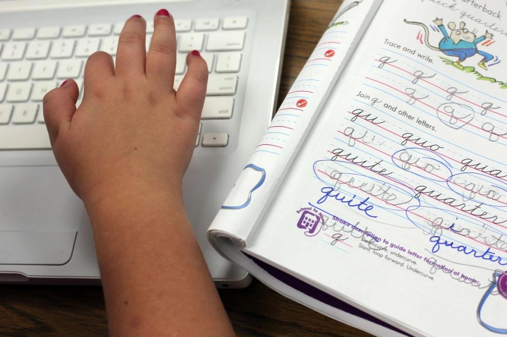 A hand typing on a keyboard next to a notebook for practicing writing in cursive
