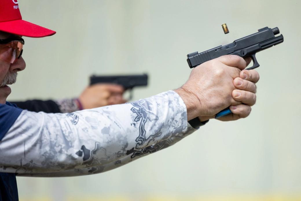 A man wearing a red hat and sunglasses points a gun at a gun range.