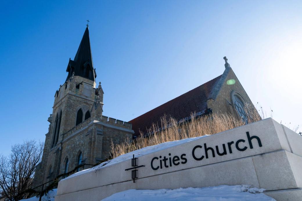 Looking upward at brick Gothic Revival style church building, modern signage in foreground that reads, "Cities Church"