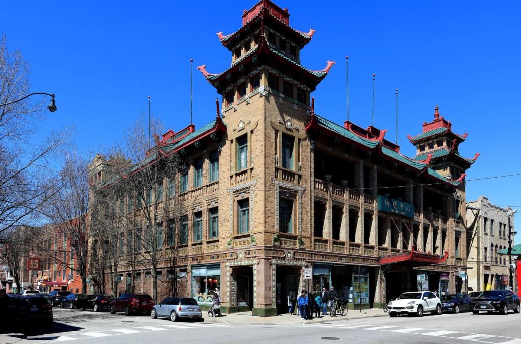 A historic Chinese-style building with ornate pagoda roofs, decorative brickwork and colorful architectural details stands on a city street, with parked cars and pedestrians in front under a clear blue sky.