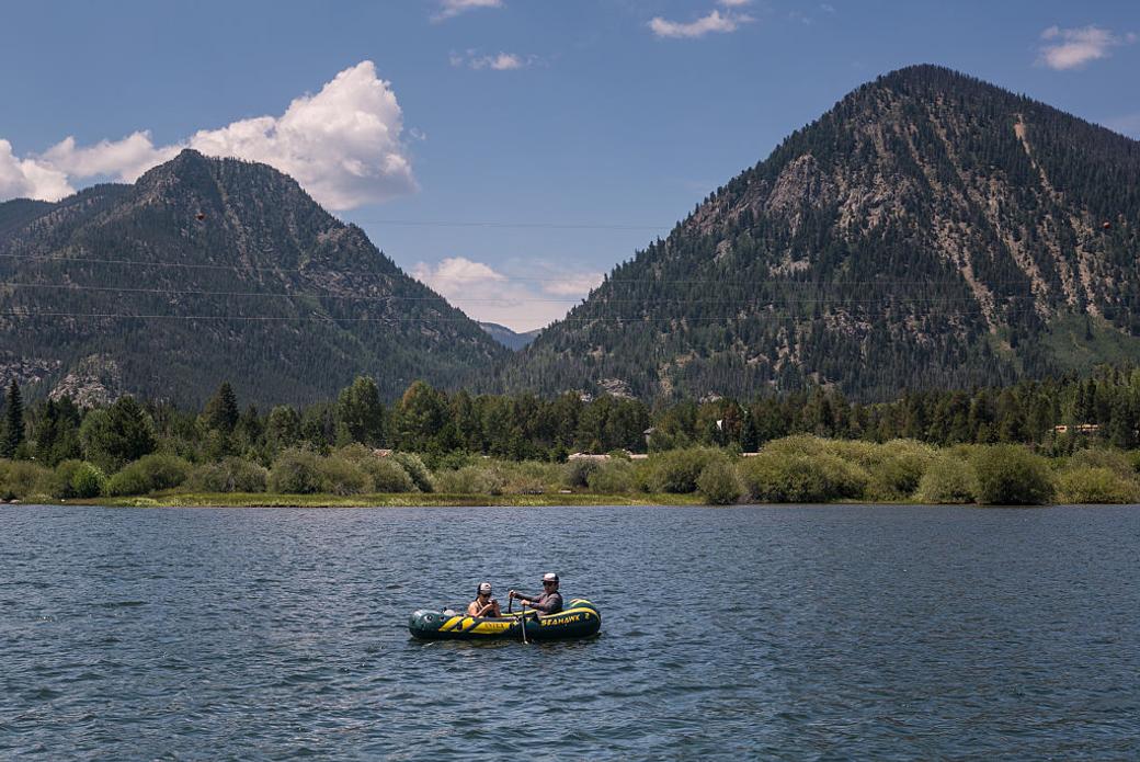 Two people on a raft in a large body of water. In the background are two hills, with trees in front of them.