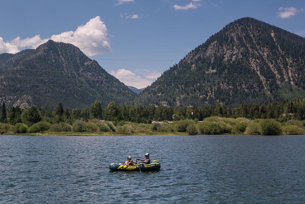Two people on a raft in a large body of water. In the background are two hills, with trees in front of them.