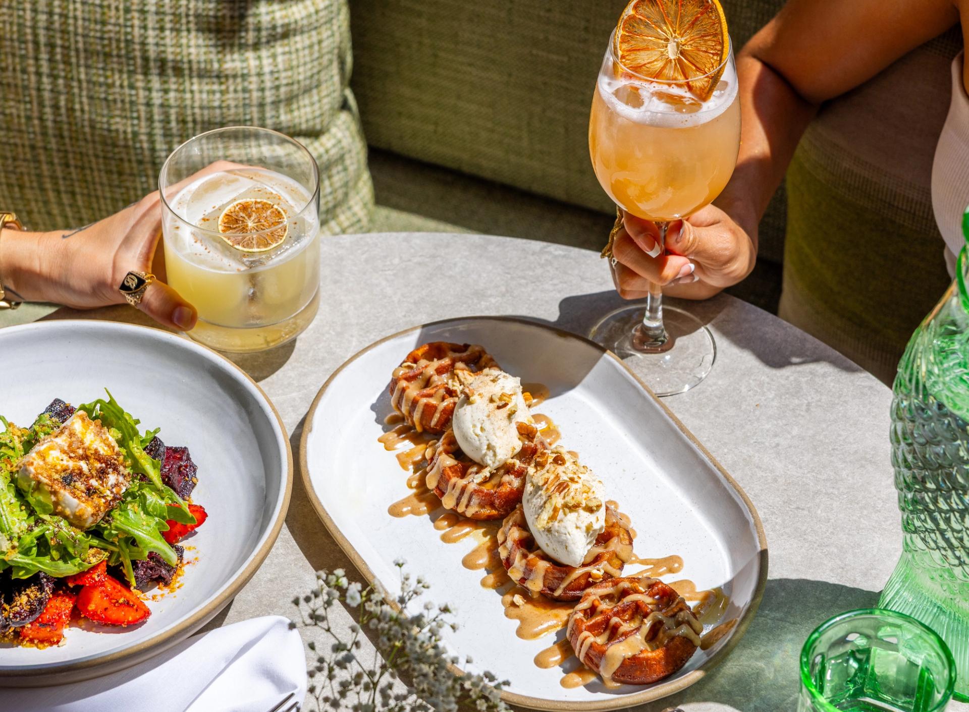 A white plate with small waffles on it, a wine glass with an orange drink in it, and a plate with salad on it.
