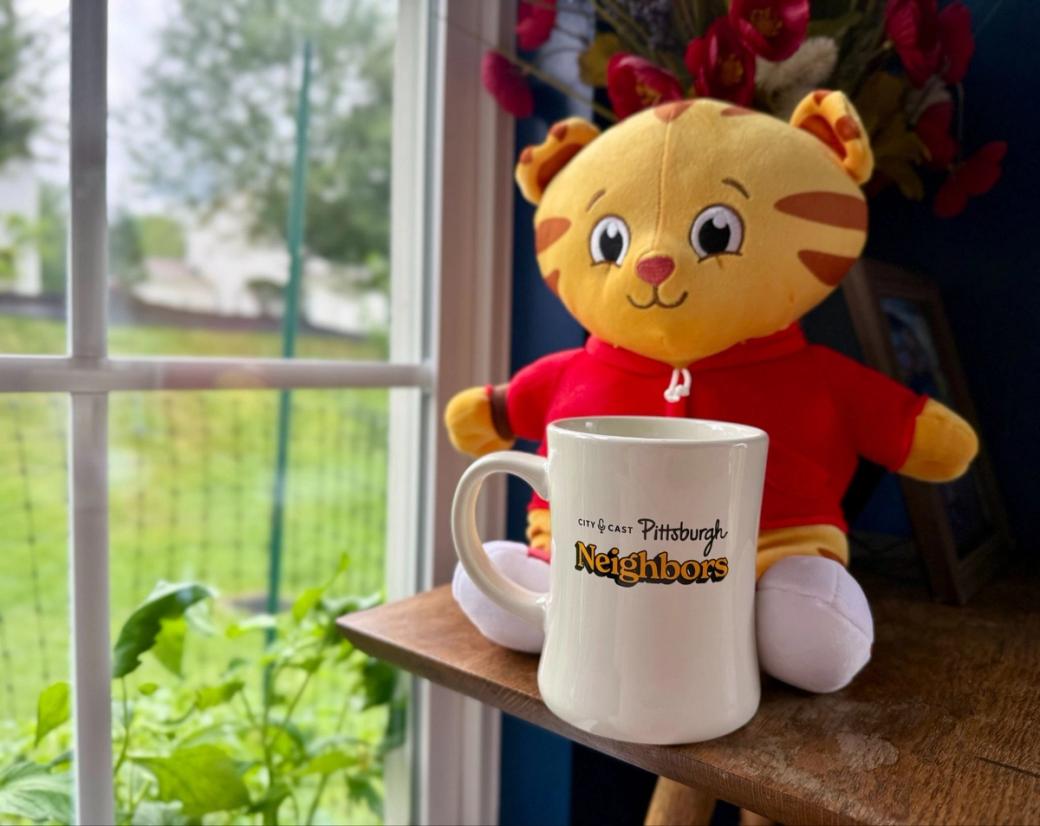 A diner-style City Cast Pittsburgh Neighbors mug sits in front of a Daniel Tiger stuffed animal.