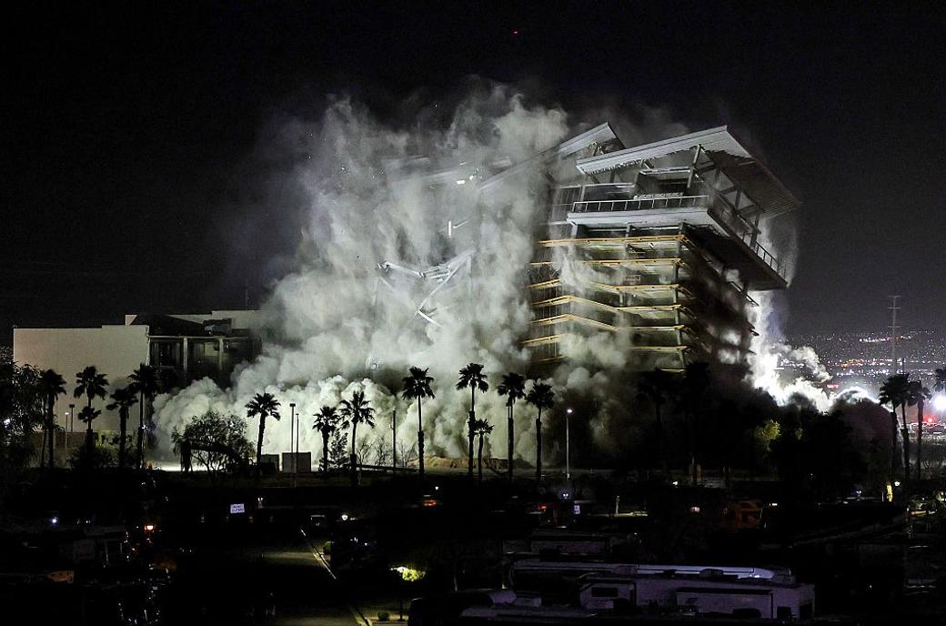 Controlled demolition of a multi-story building at night, collapsing into massive clouds of dust and smoke behind a row of palm trees.