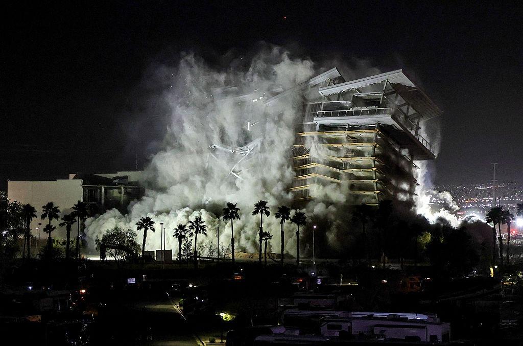 Controlled demolition of a multi-story building at night, collapsing into massive clouds of dust and smoke behind a row of palm trees.