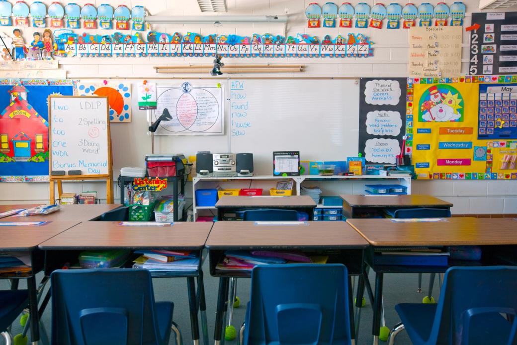 Desks sit empty in a kindergarten classroom with walls covered in colorful posters. 