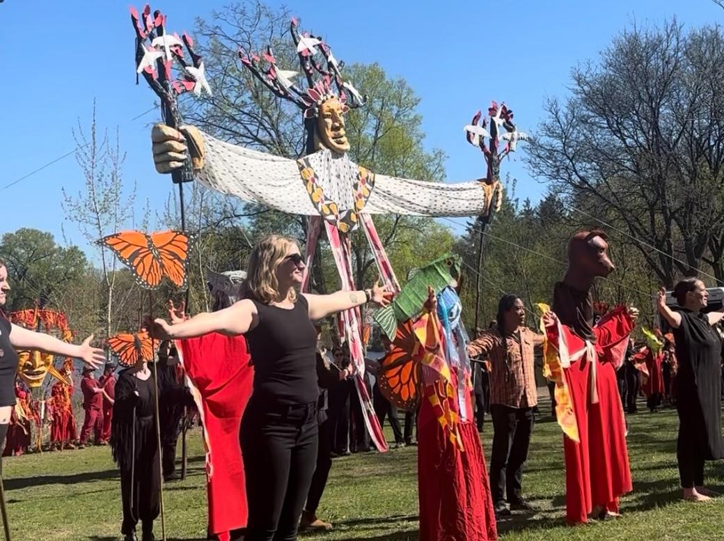 Group of people with arms outstretched stand in group around large maypole puppet outdoors