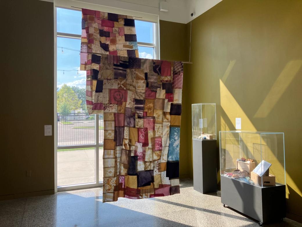 A sewn-together cloth of smaller pieces of cloth dyed in shades of pinks, yellow, purple, and blue, hand from the ceiling on display. To the right are two display cases filled with yarn and cotton.