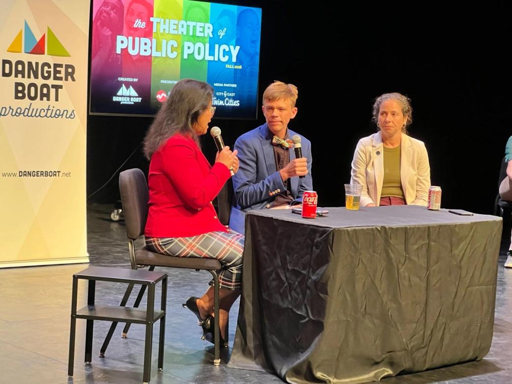 Two women and a man sit behind a table on a stage. The woman on the left and the man in the middle are holding microphones. There is a screen behind them that reads, "the THEATER of PUBLIC POLICY."
