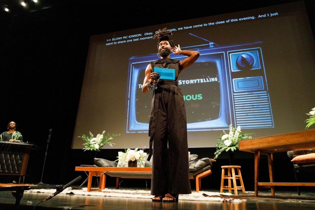 Elijah McKinnon standing on a stage at the Museum of Contemporary Art Chicago with a projector screen in the back showing an old-school TV.