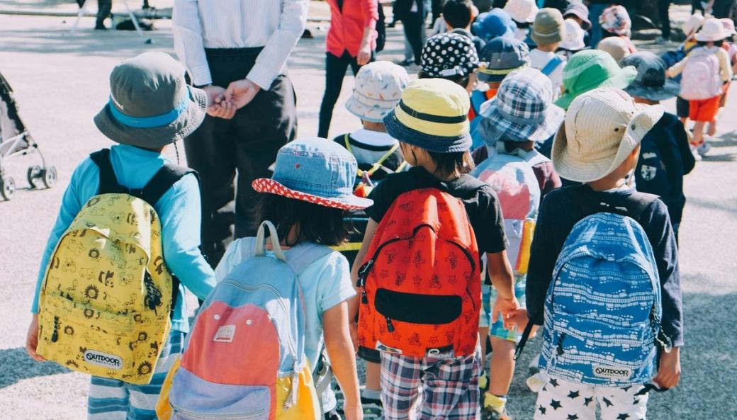 A crowd of young students in brightly colored hats and backpacks hold hands while walking down the sidewalk.