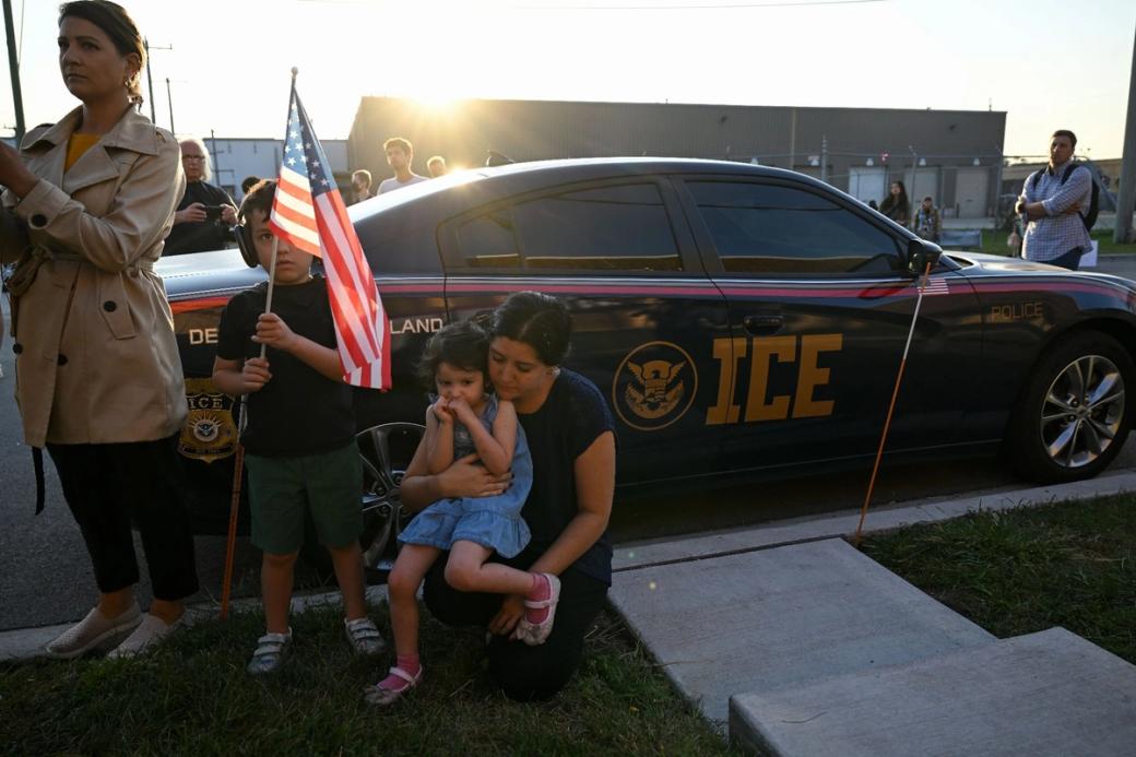 Two women and two children standing in front of a car that says "ICE" on it. One of the children is sitting in one of the woman's lap and the other child is holding a US flag