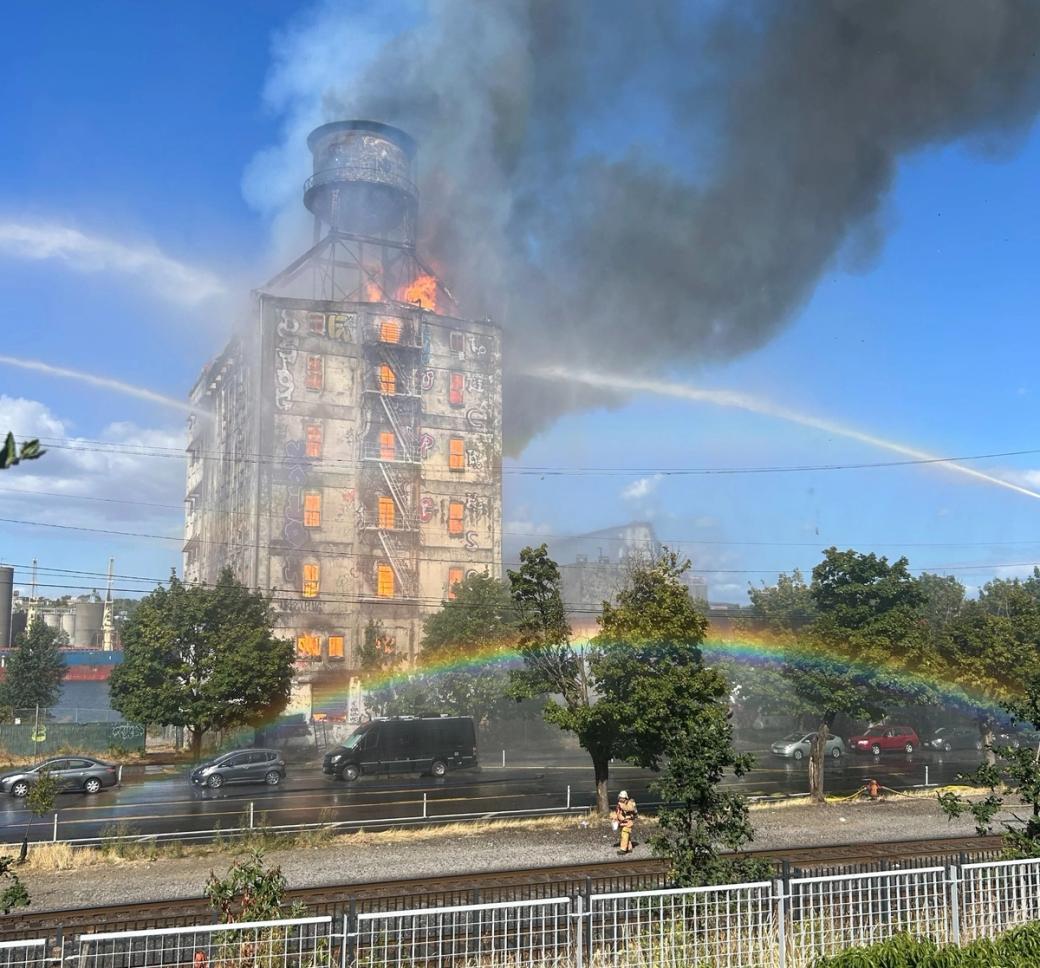 A tall, graffiti-covered building with flames inside and dark smoke bellowing out the top, multiple fire-hose streams targeting the building and a rainbow, trees, and cars in the foreground.   