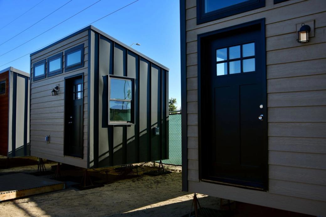 A few tiny homes in a row. The complete front and side of one of them is visible. They have beige-colored walls in the front and a dark-colored door, plus three windows at the top and one on the side.