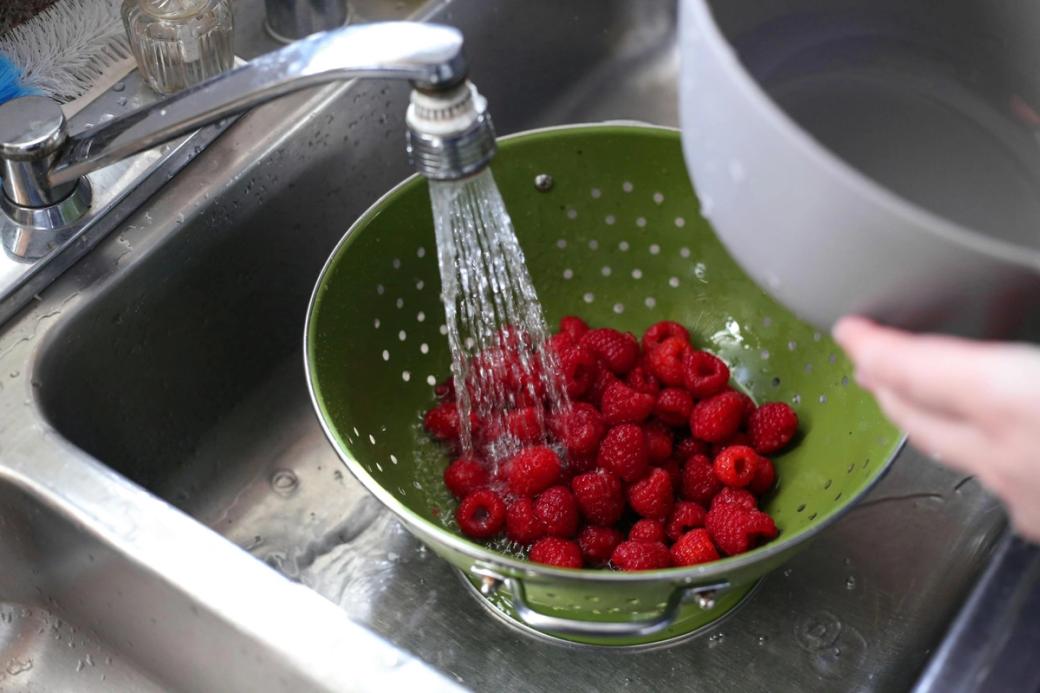 Raspberries in a green colander in the sink under running water.