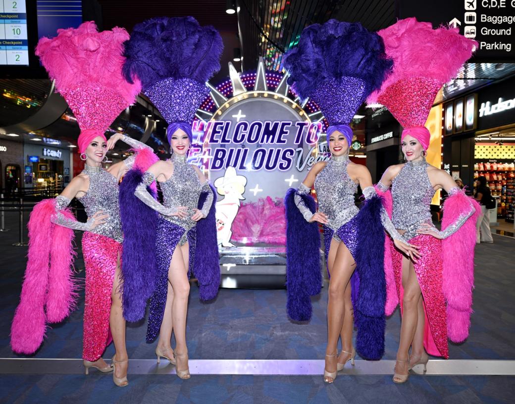 Showgirls at the airport in front of a sign. 