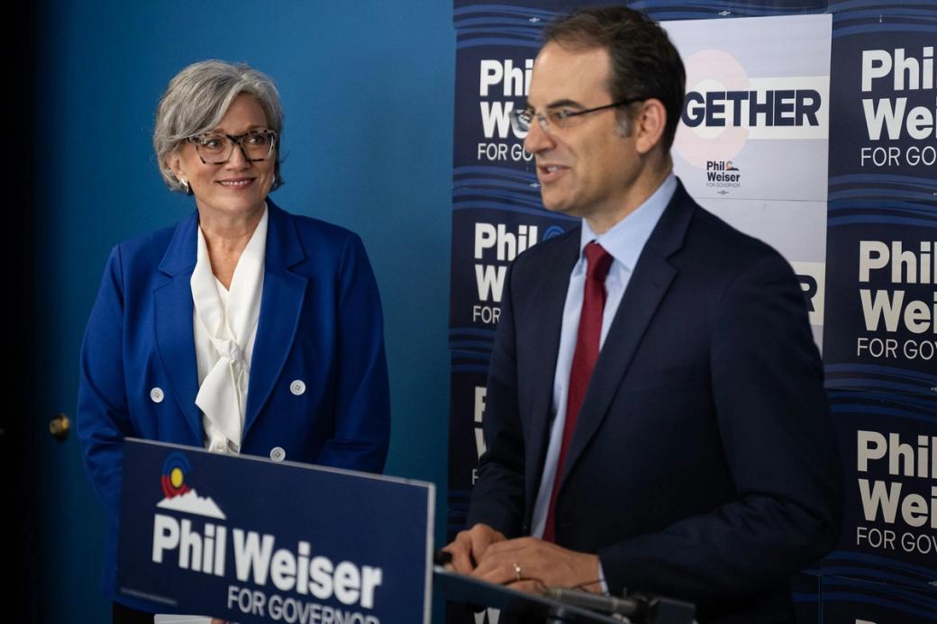  A man in a suit standing at a podium, with "Phil Weiser for Governor" behind him and on the podium. A woman in a blue coat is standing next to him.