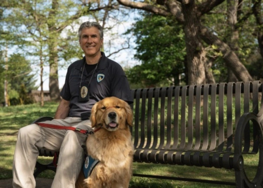 A police officer sits on a bench next to a golden retriever.